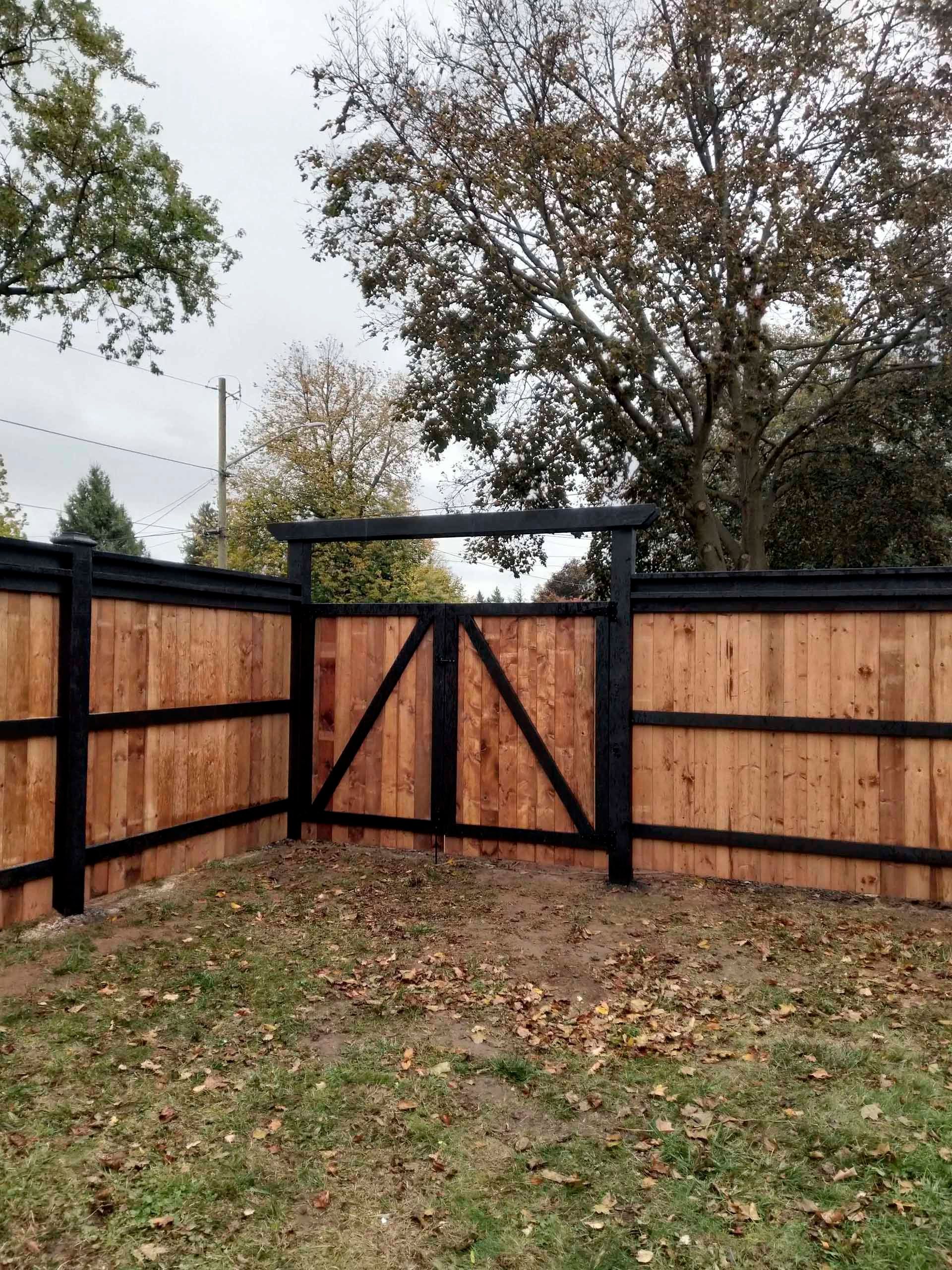 Wooden fence and gate with black trim and natural wood panels, in a grassy yard, under a cloudy sky.