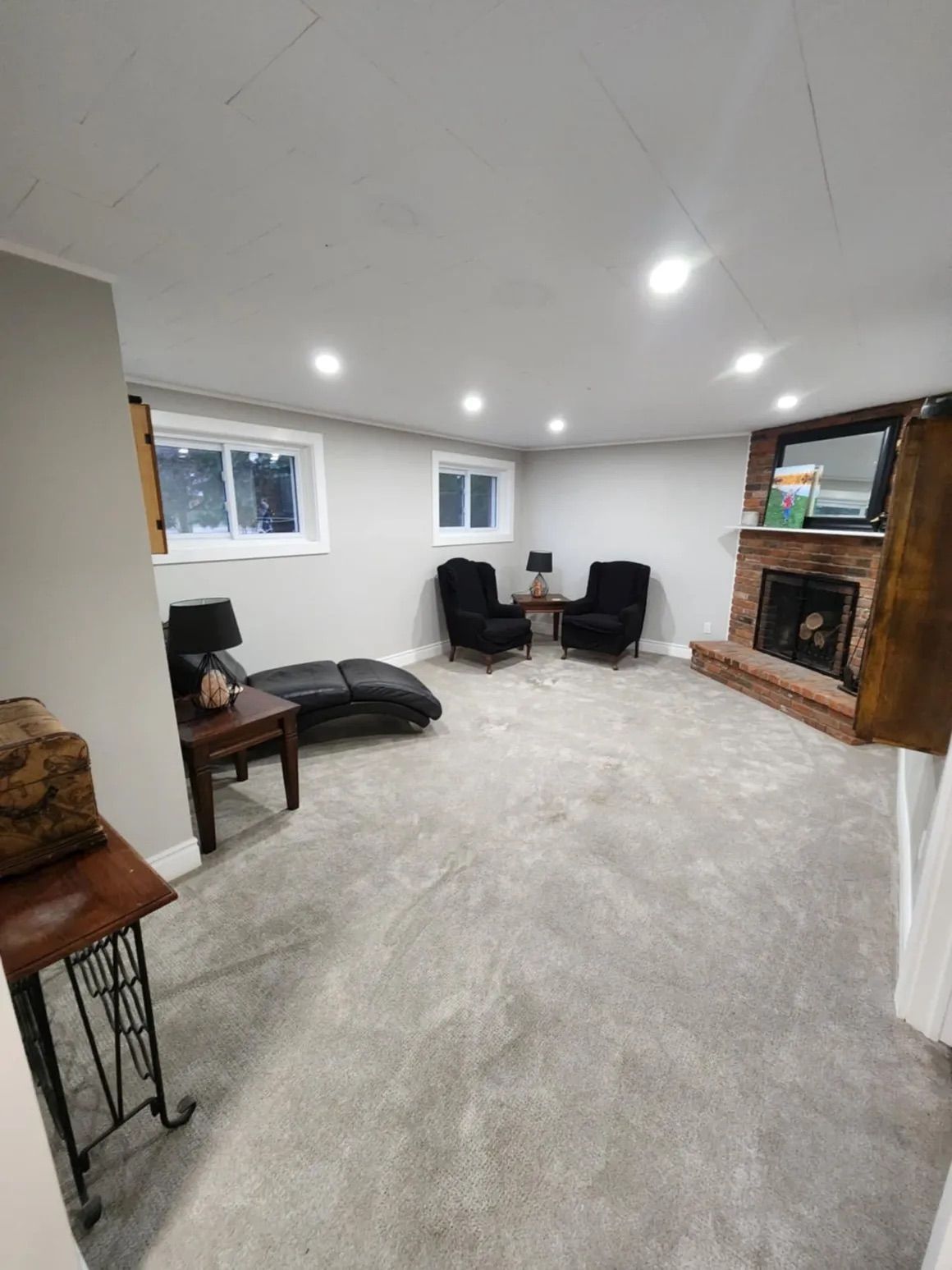 Cozy basement room with gray carpet, two black armchairs, chaise lounge, fireplace, and small windows.