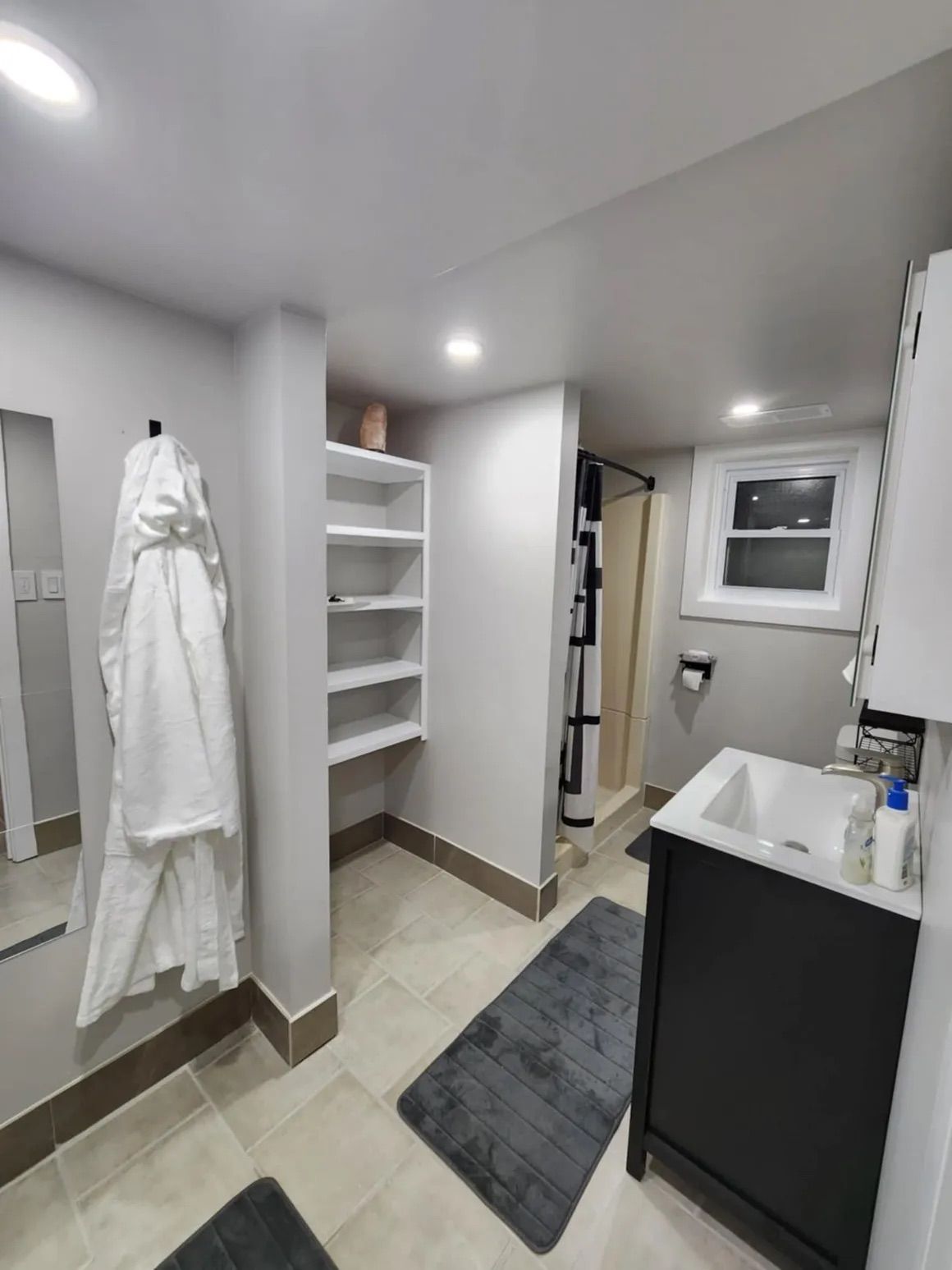 Small, modern bathroom with neutral tones. White robe hangs by a mirror; vanity and shower in view.