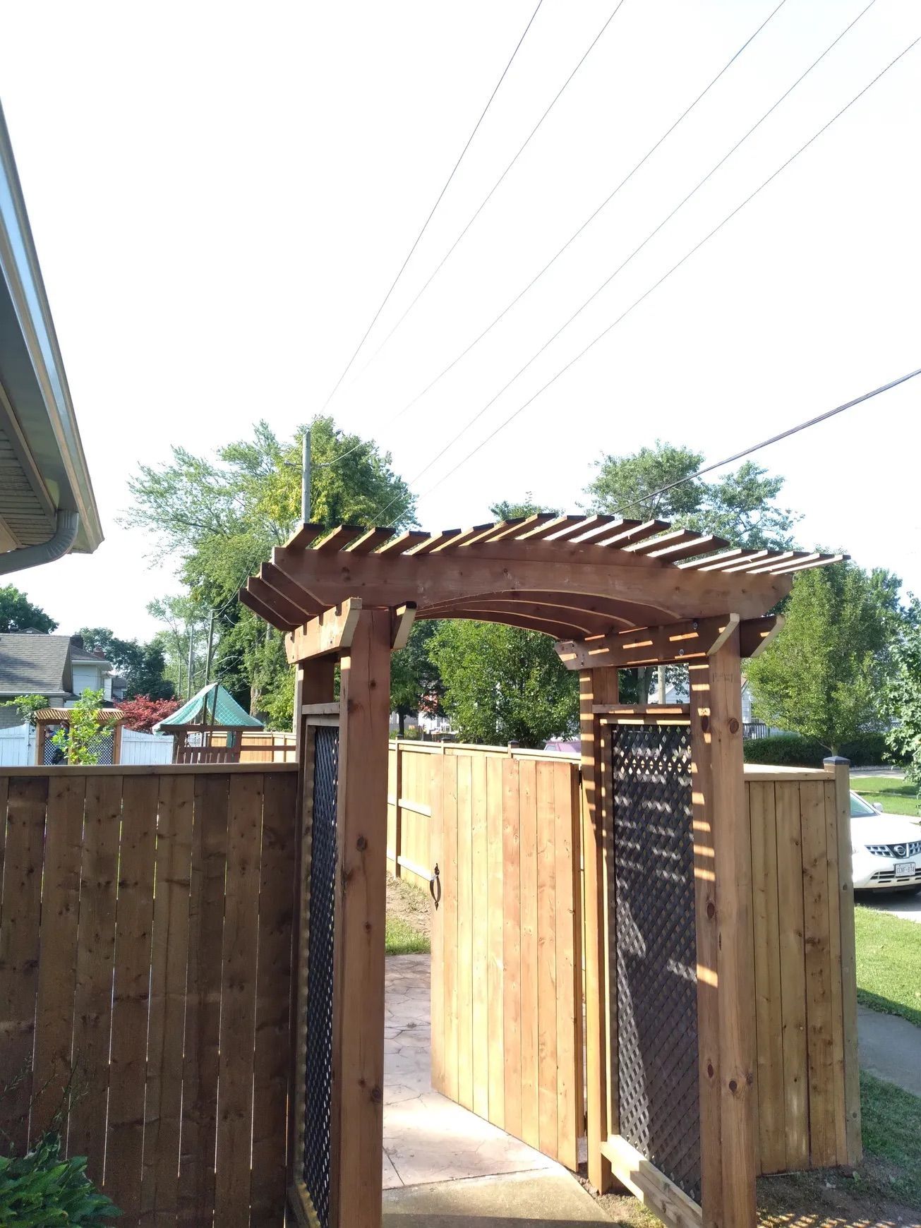 Wooden arbor and fence, leading to a gate. Sunny day, residential setting.