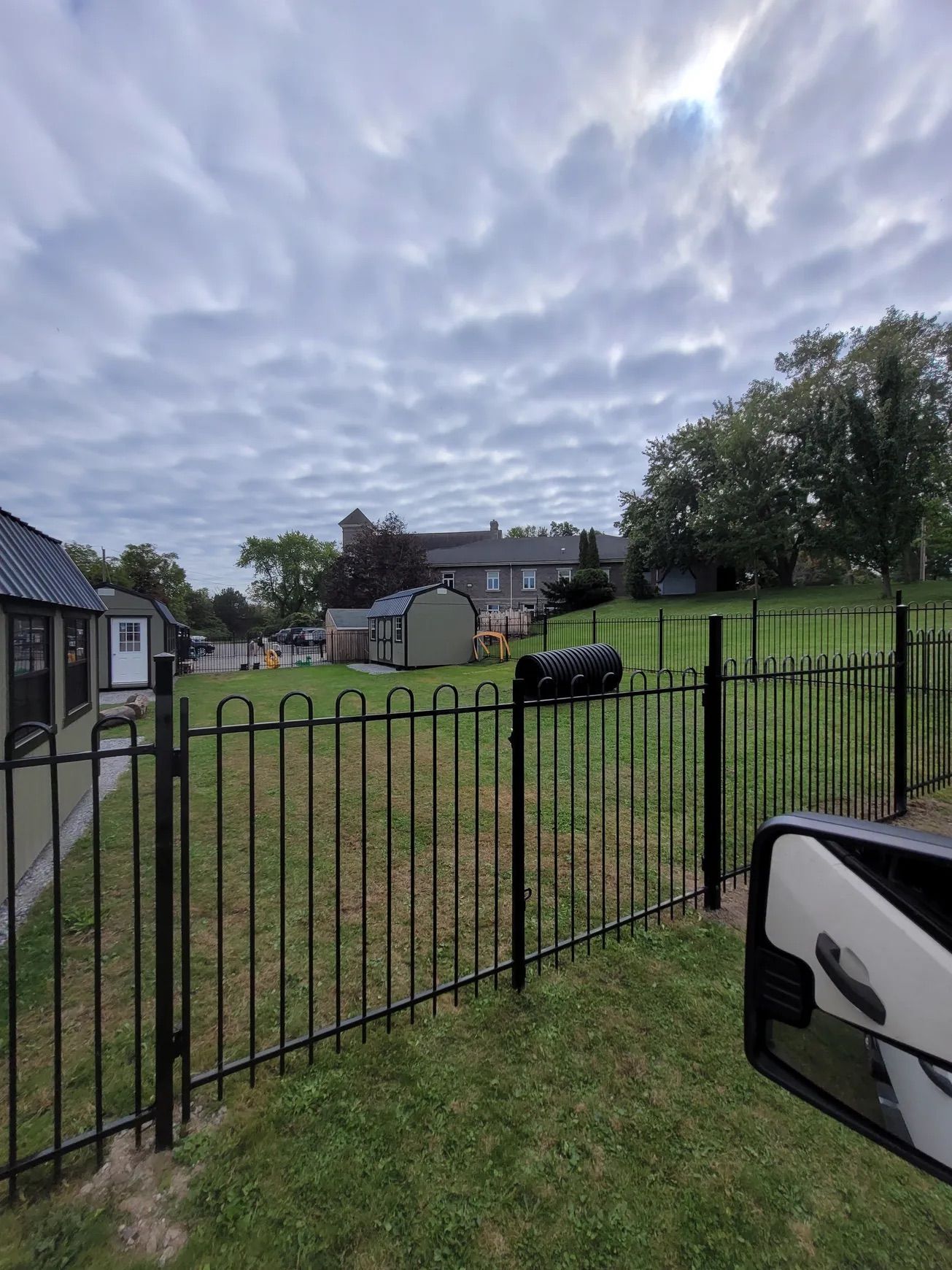 Black metal fence bordering a grassy area with buildings and cloudy sky in the background.