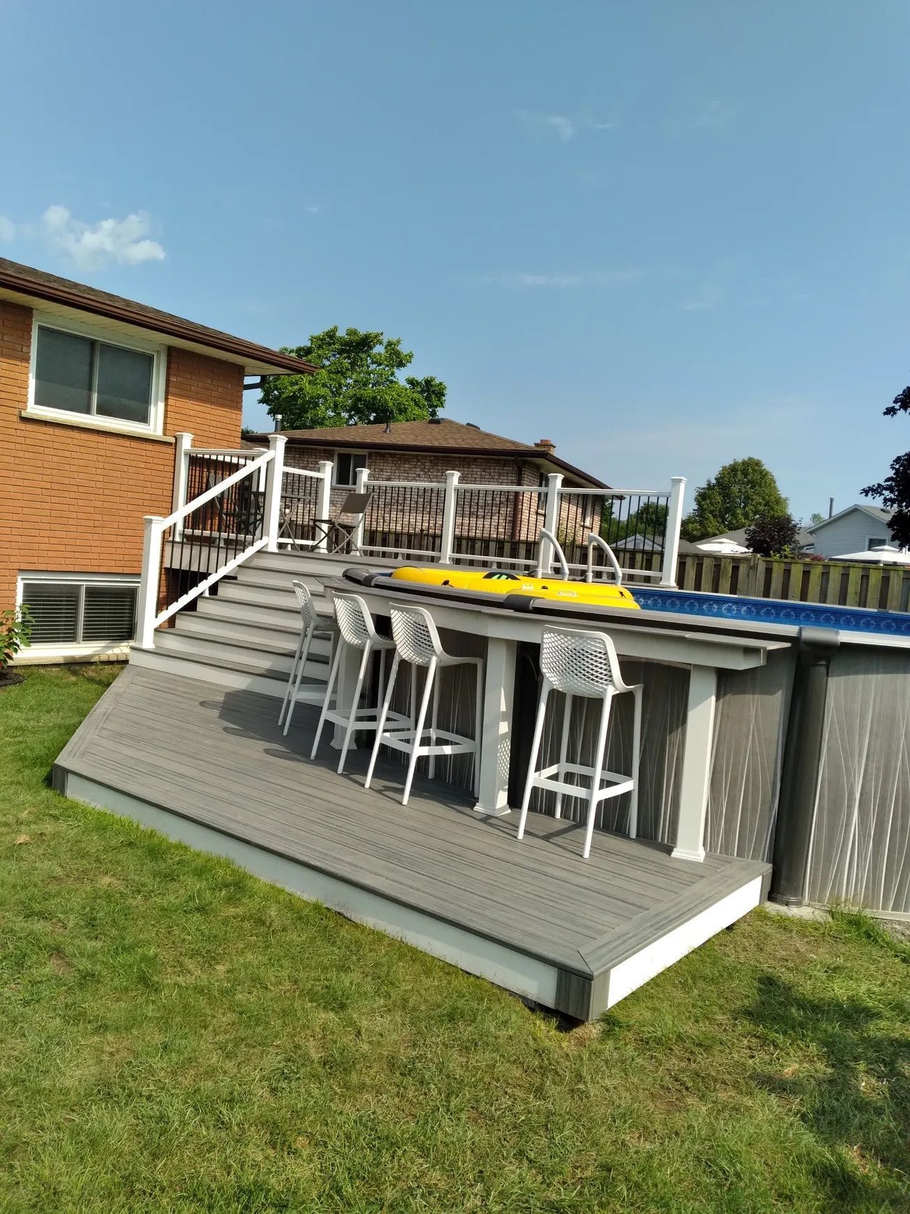 Backyard with a pool deck, white bar stools, and a house in the background. Green grass and blue sky.