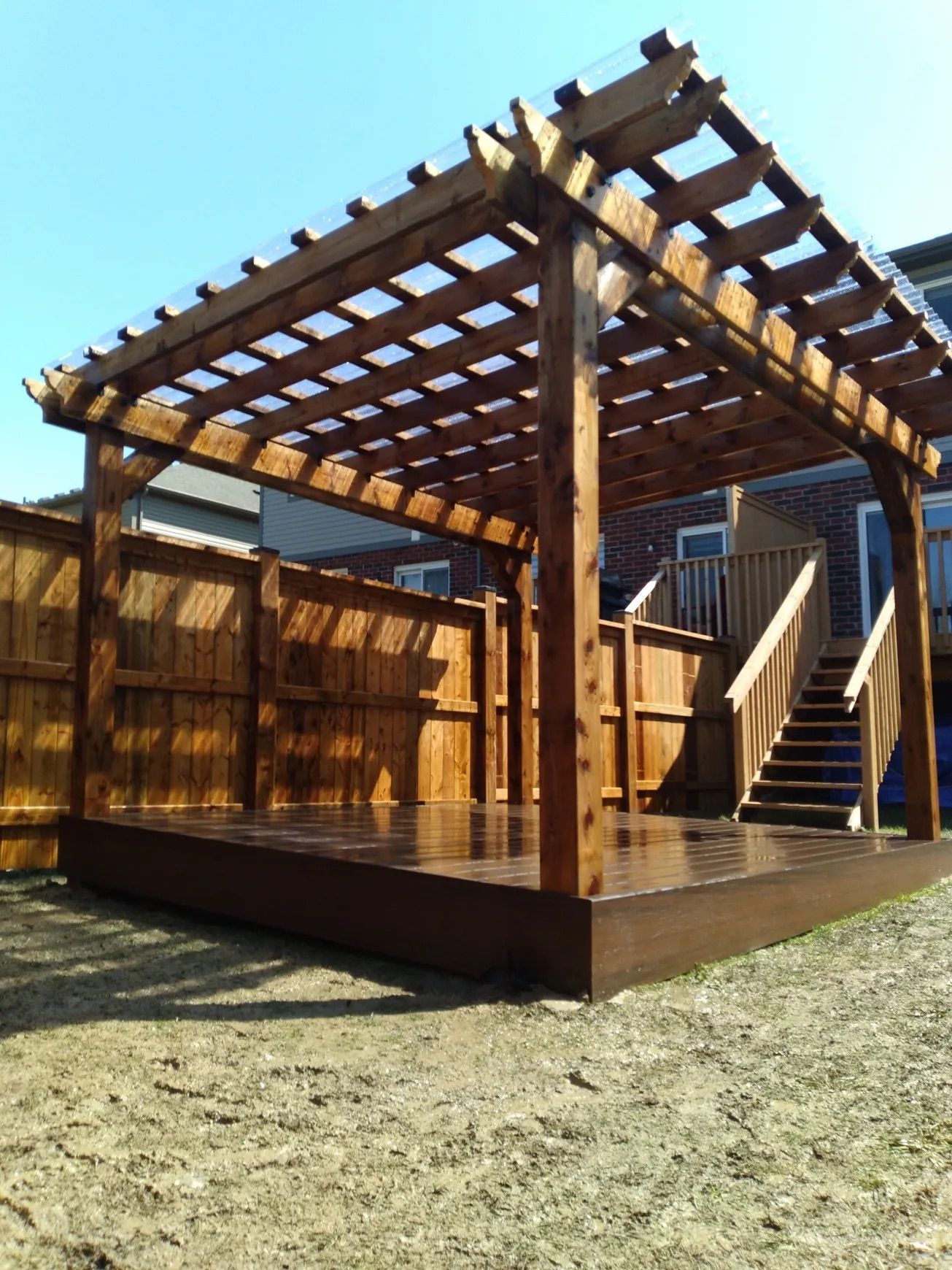 Wooden pergola on a brown deck, with stairs. Fence in the background.