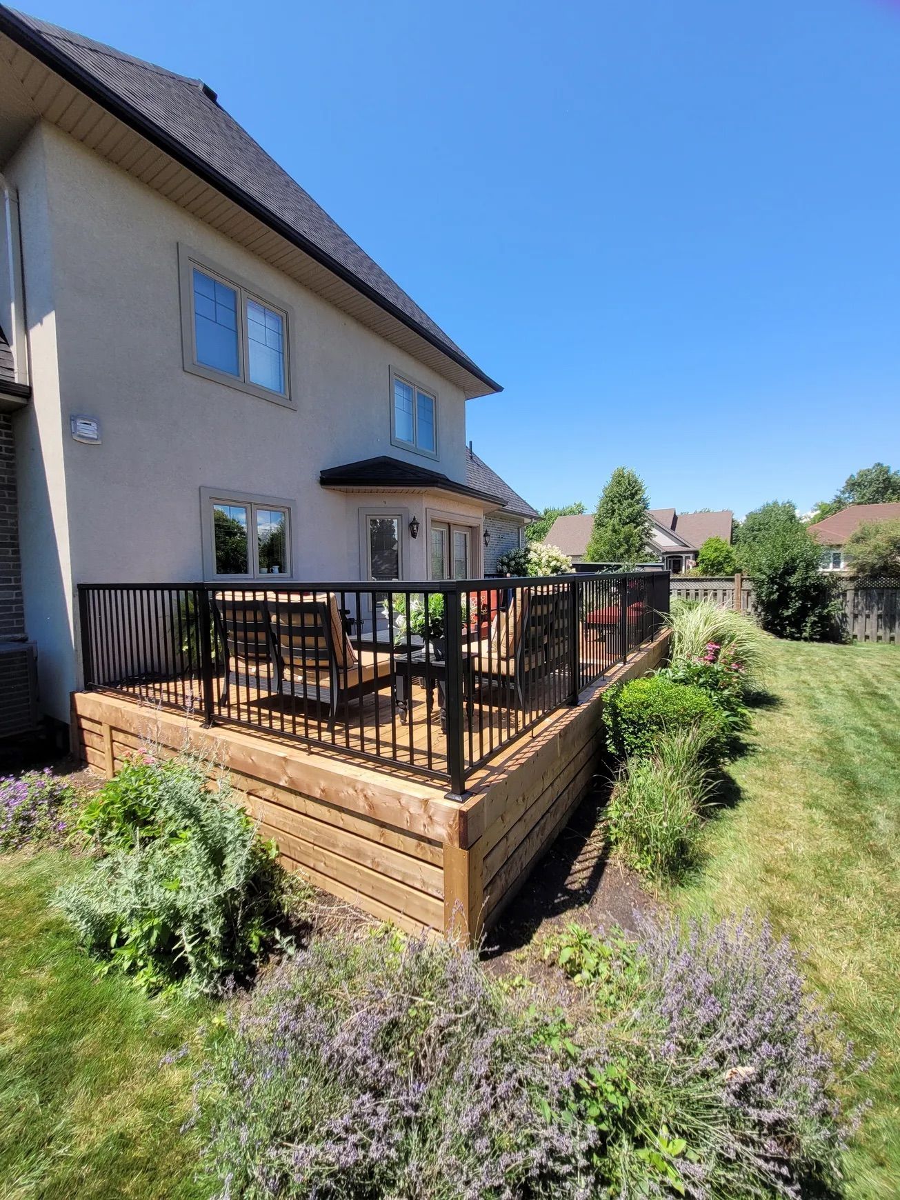 Wooden deck with black railings, attached to a beige house, under a blue sky.