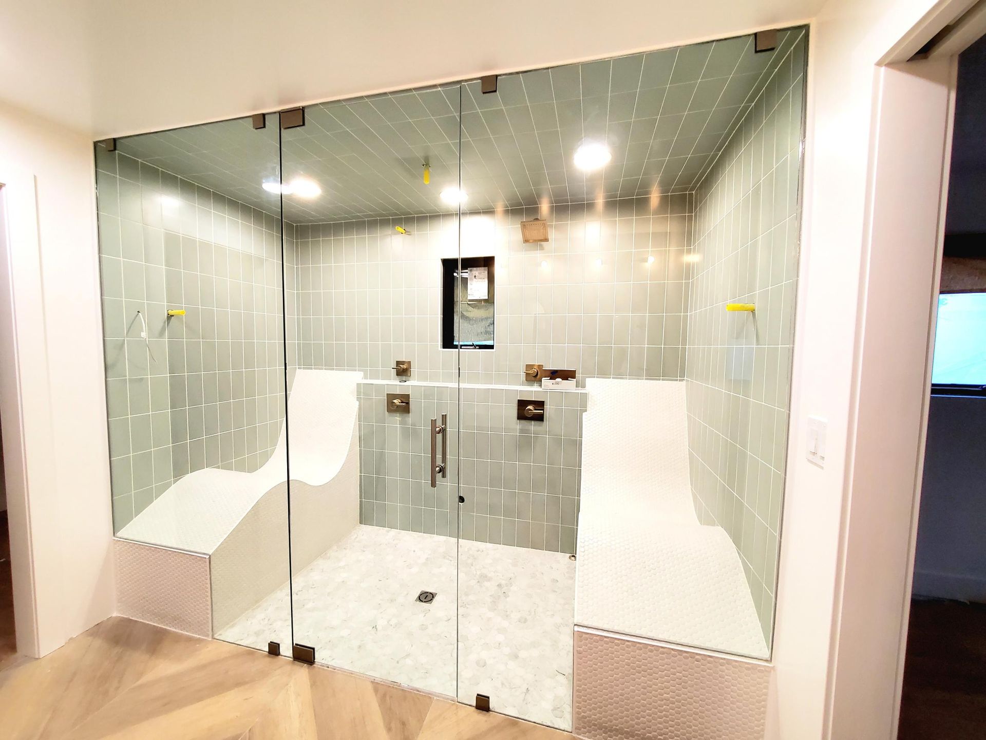 Glass-walled, tiled shower with built-in white benches. Light wood floor outside the shower.
