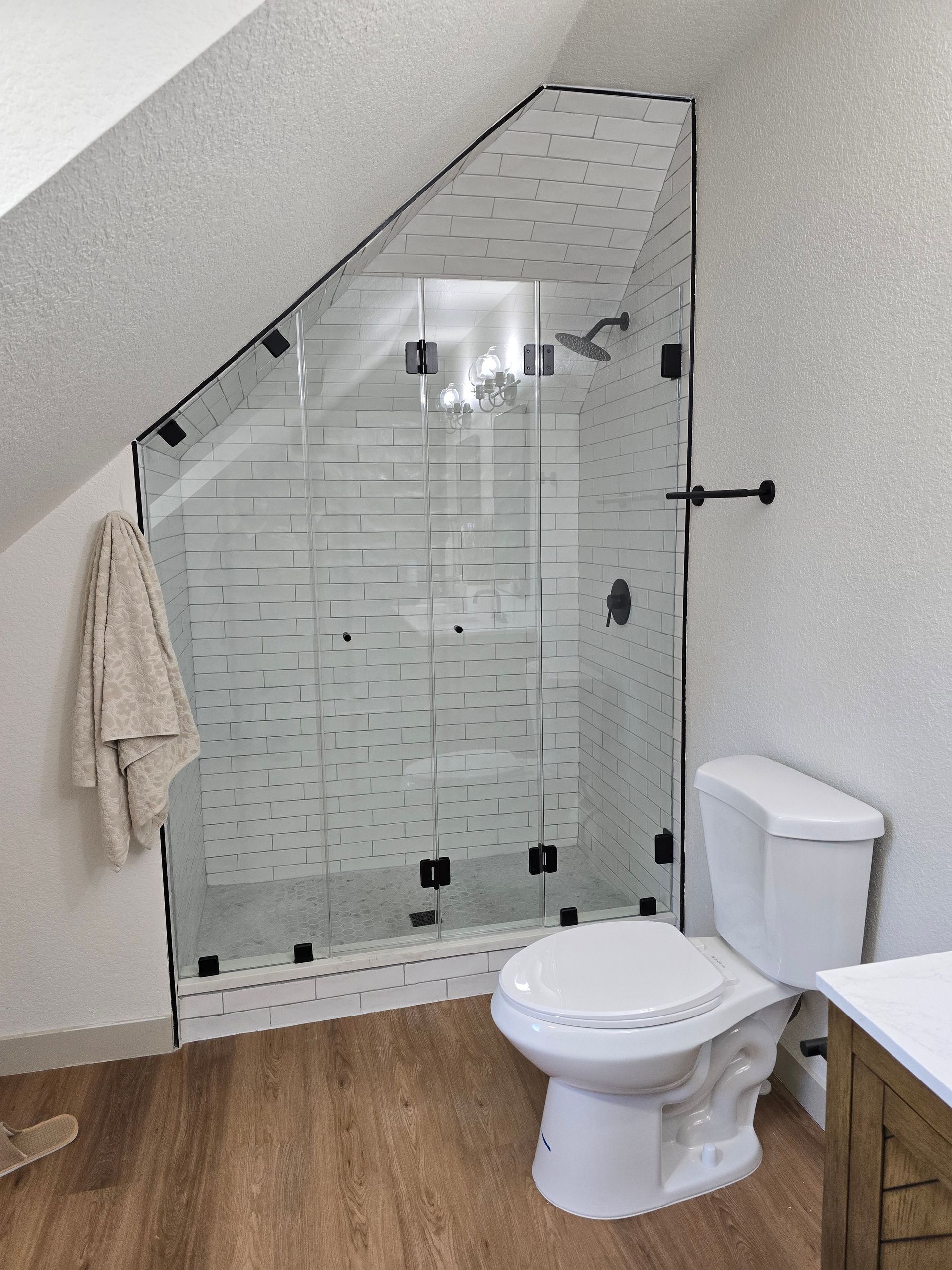 Bathroom with a sloped ceiling featuring a glass shower, white toilet, wooden floors and textured walls.