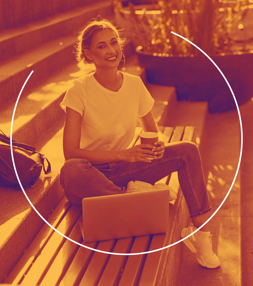 Uma mulher está sentada em um banco com um laptop e uma xícara de café.