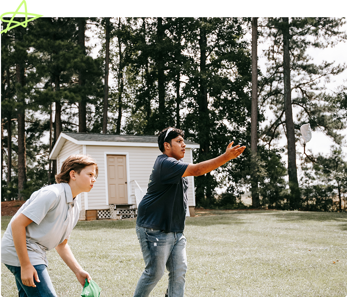 Two people outdoors playing catch in a grassy yard beside a small shed and tall trees.