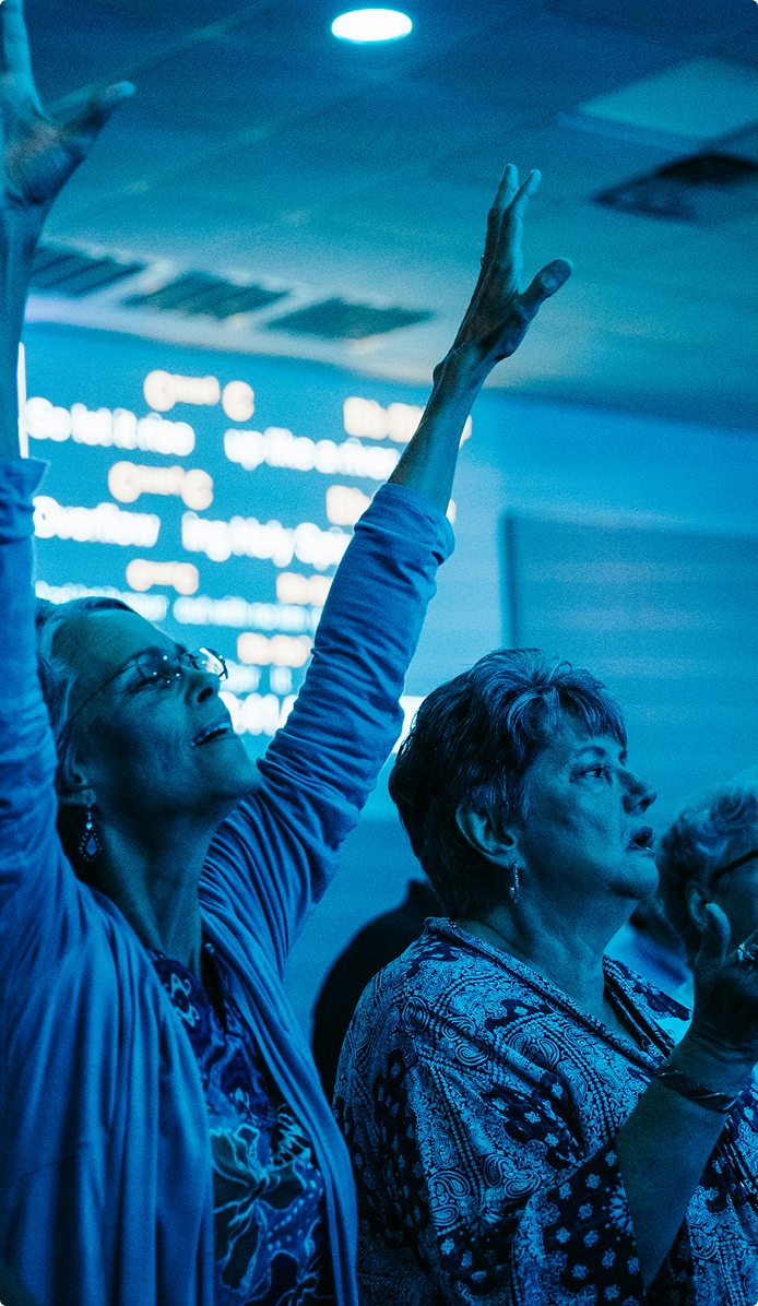 Audience at a blue-lit event, one person raising an arm toward a bright screen in the background