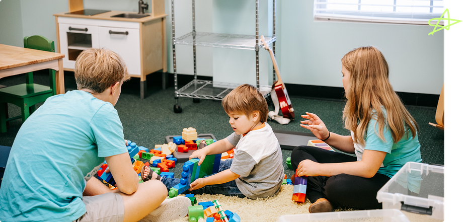 Three children sit on the floor playing with toys in a classroom or daycare room.