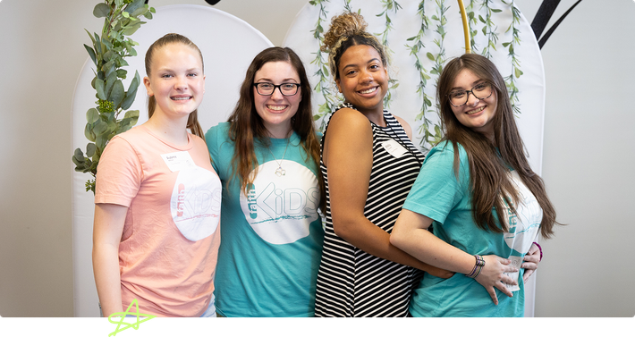 Four women smiling and posing together in front of a white and green floral backdrop.