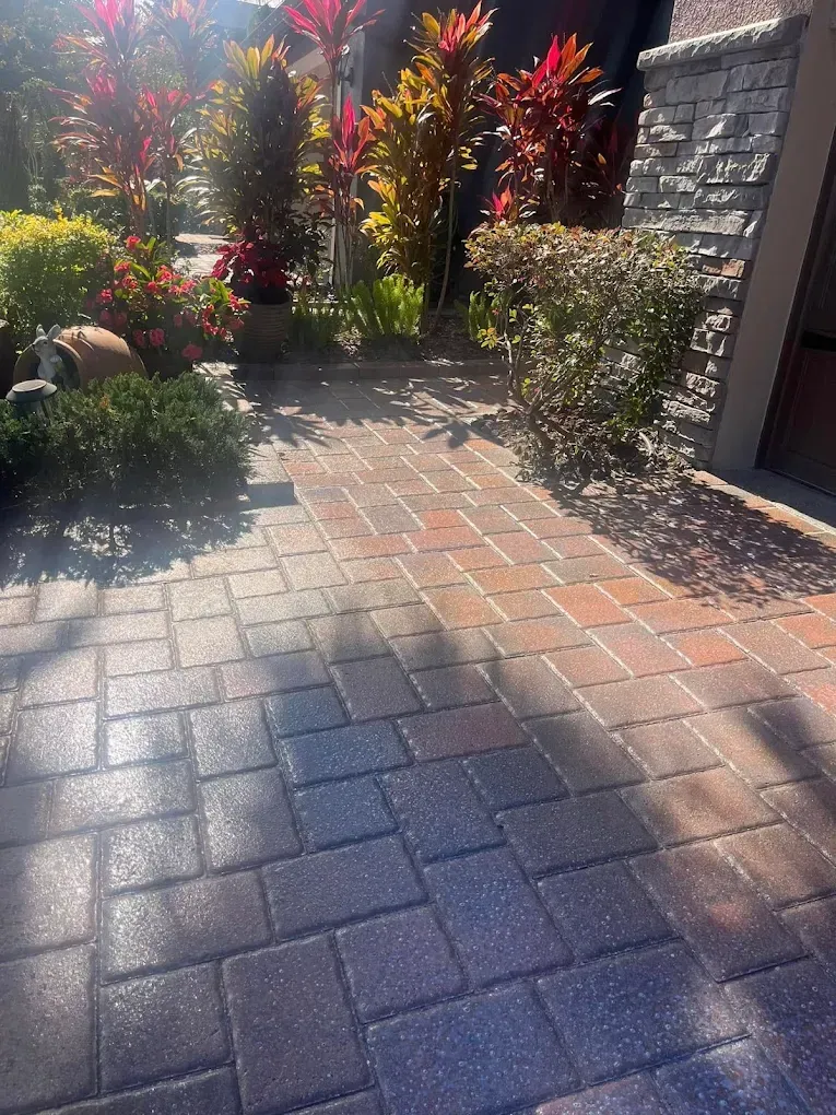 A brick walkway leading to a house with flowers in the background.