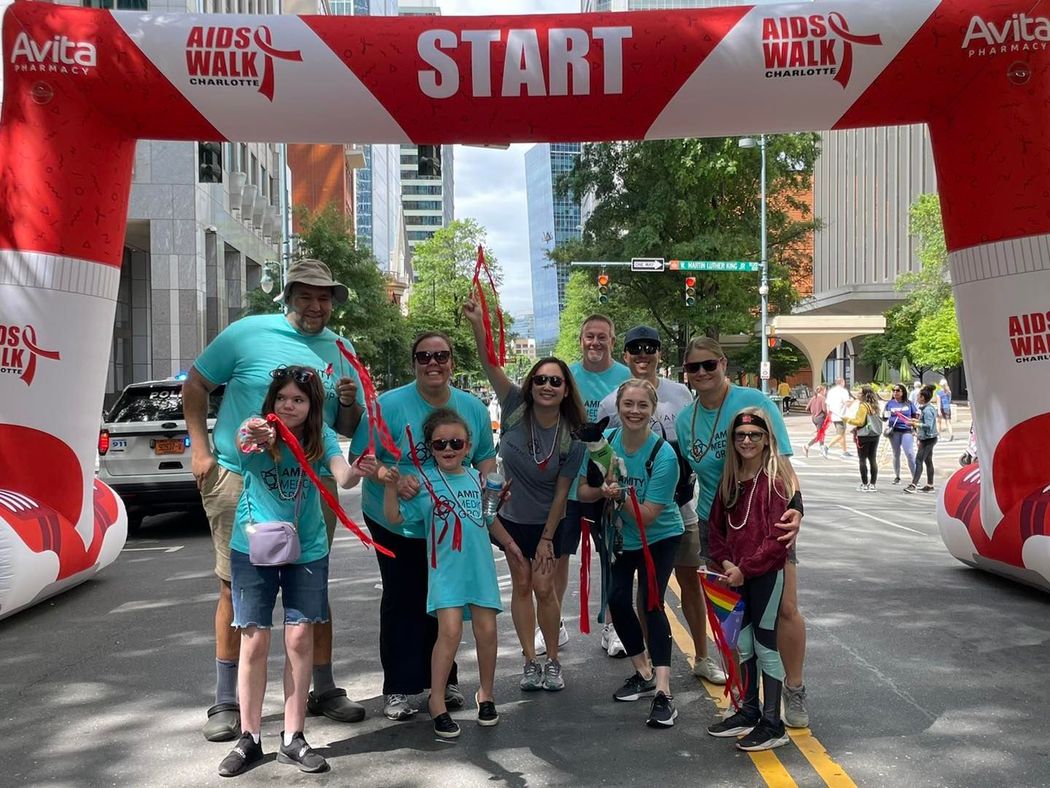 Group of people smiling at the AIDS Walk starting line, under a red and white archway.