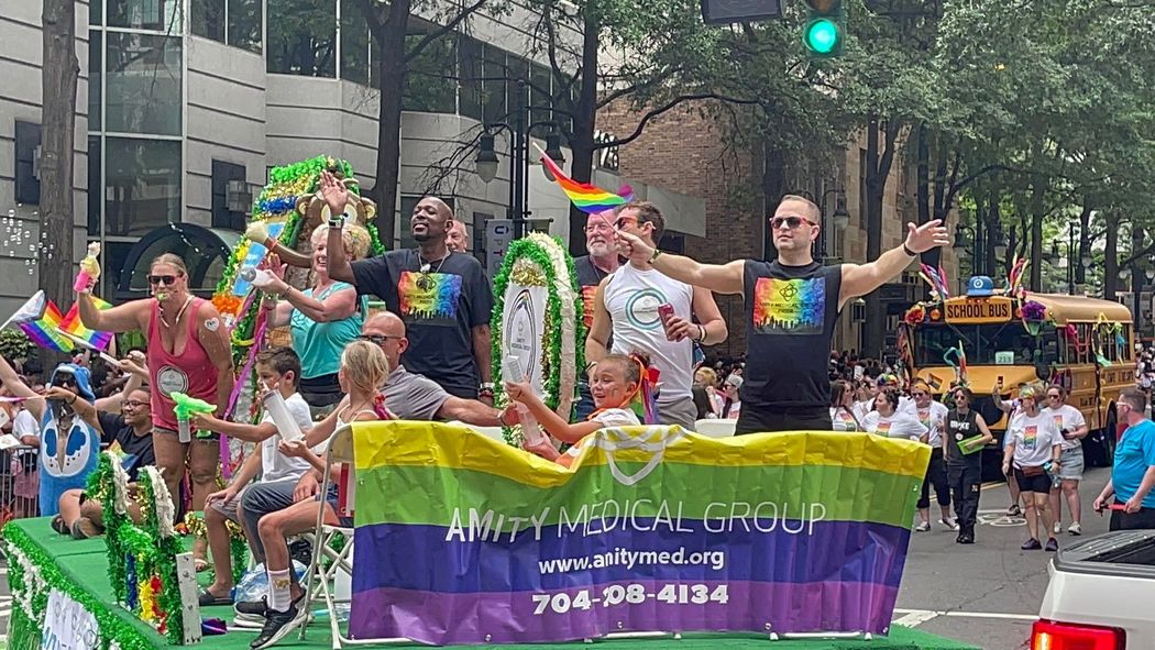 People on a float at a Pride parade, waving arms, wearing rainbow colors, with banner 