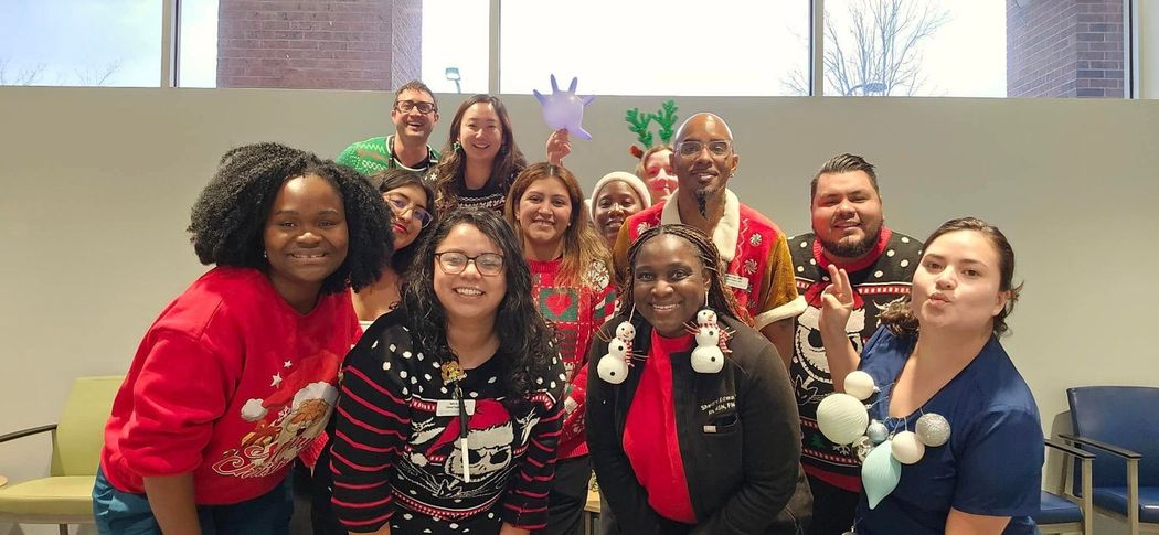 Group of people in holiday sweaters posing, smiling. Festive office setting with brick wall.