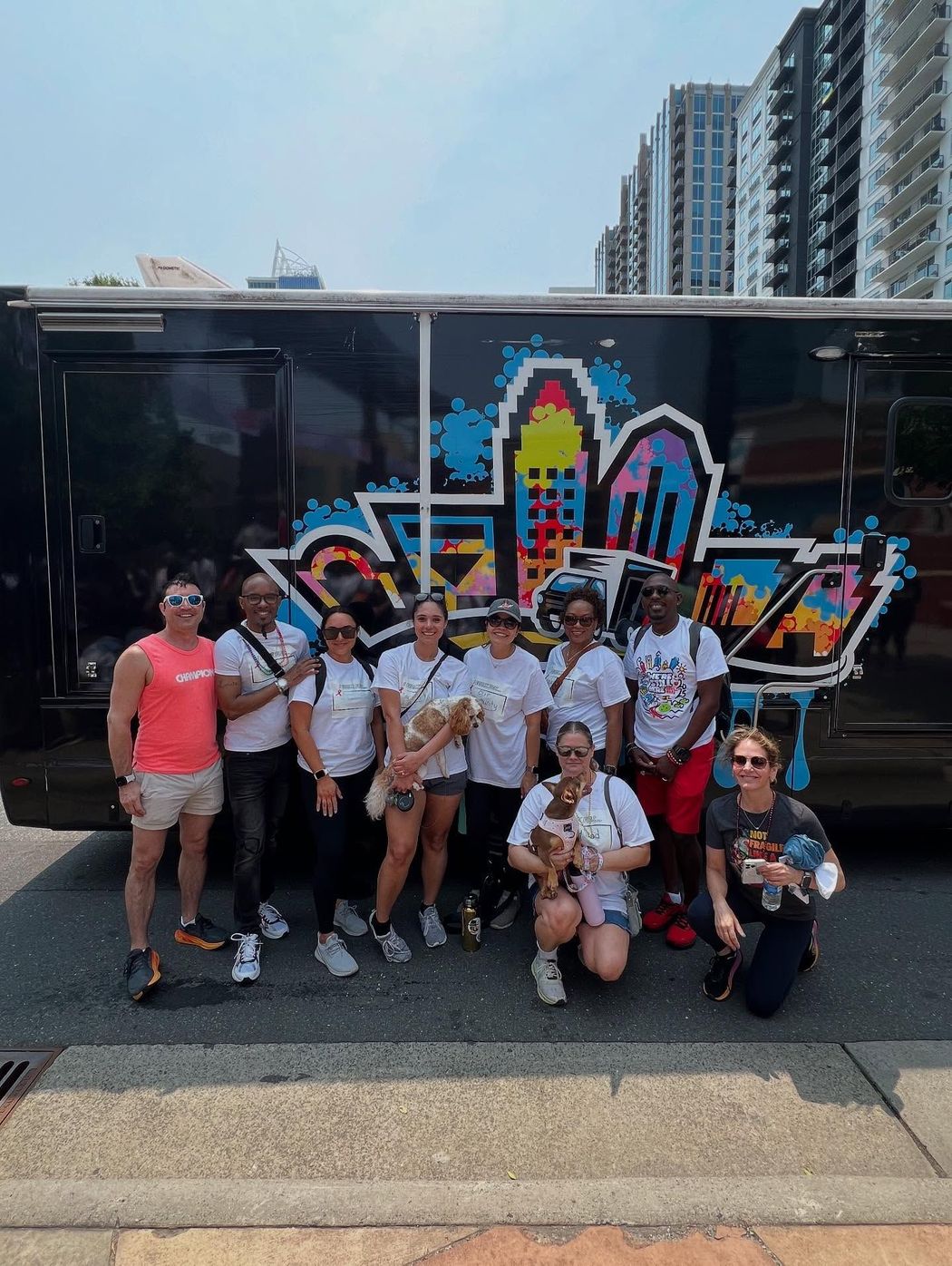 Group of people posing with dogs in front of a black bus with city-themed graffiti.