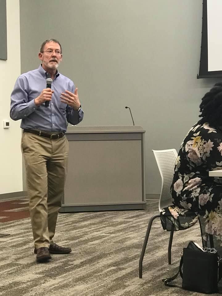 Man in blue shirt and khaki pants speaks at a podium, gesturing with his hands. Woman in floral print seated.