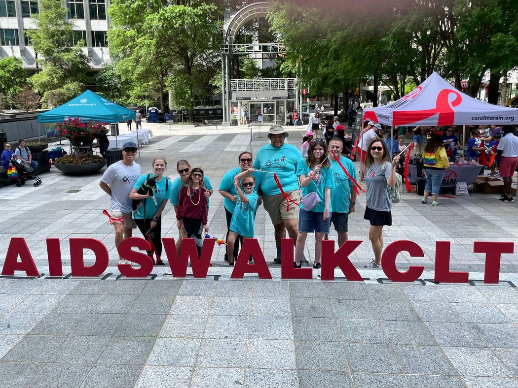 People in teal shirts at AIDS Walk CLT, red letters on ground. Outdoor event with tents.