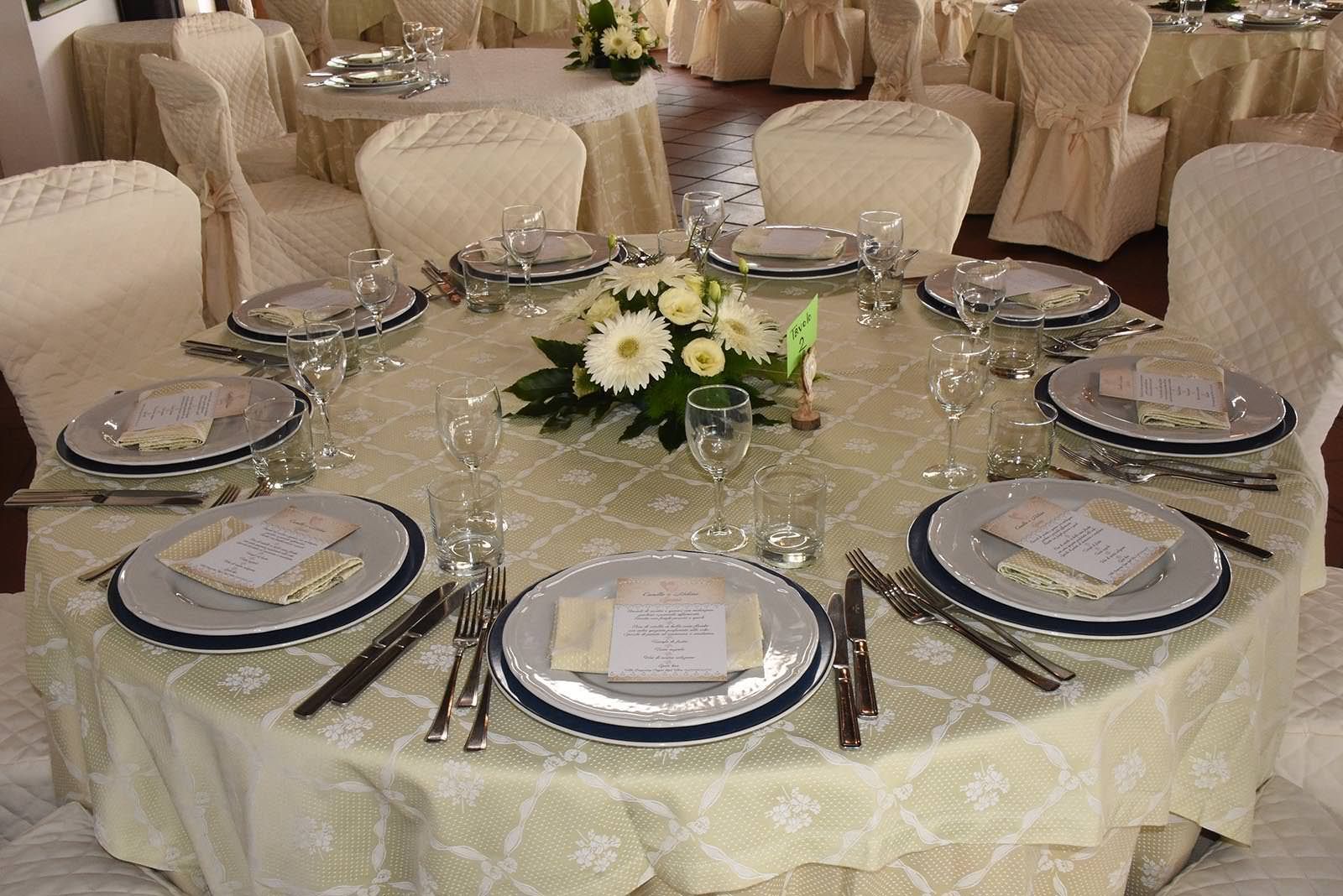 Tavolo rotondo apparecchiato per una cena formale, decorato con segnaposto e un centrotavola floreale.