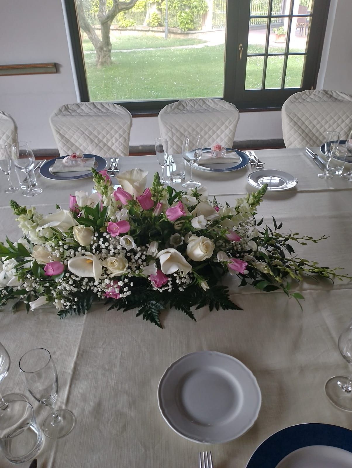 Lungo tavolo apparecchiato per un pasto con centrotavola floreale. Tovaglie, posate e sedie bianche visibili vicino a una finestra.