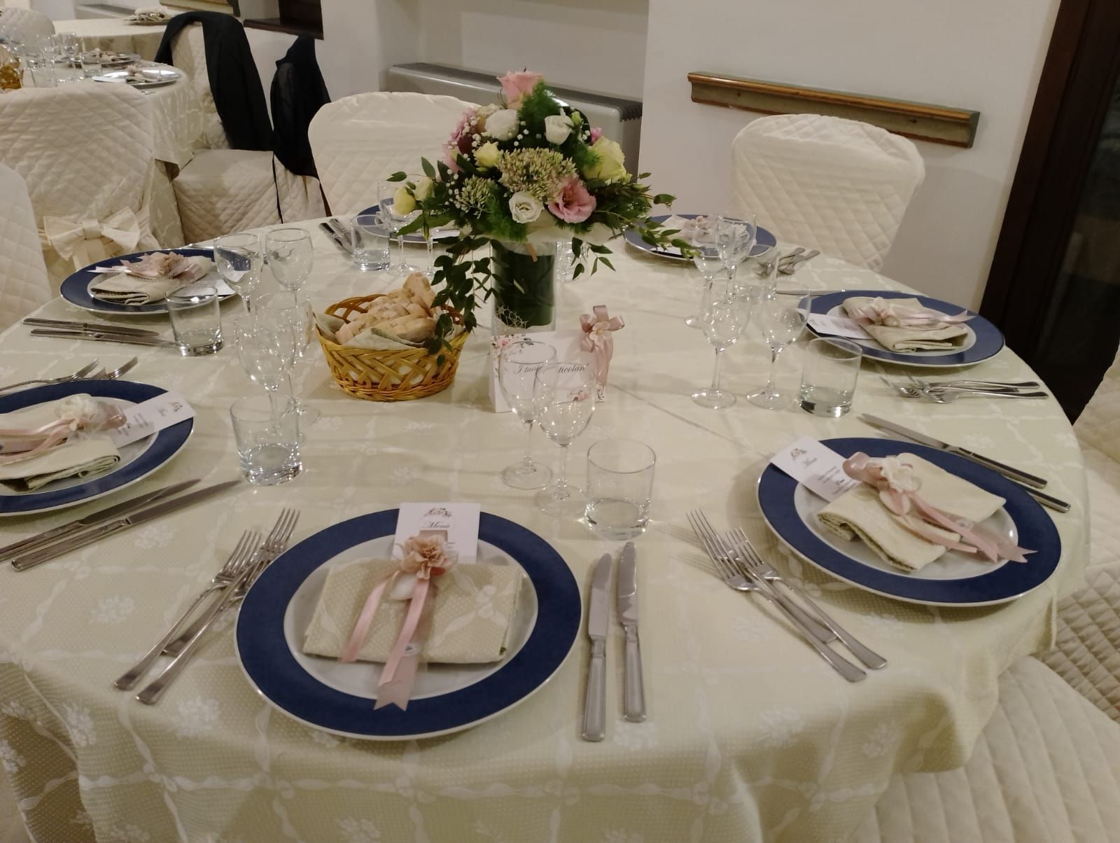 Tavolo rotondo apparecchiato per un pasto, con piatti, posate e centrotavola floreale.