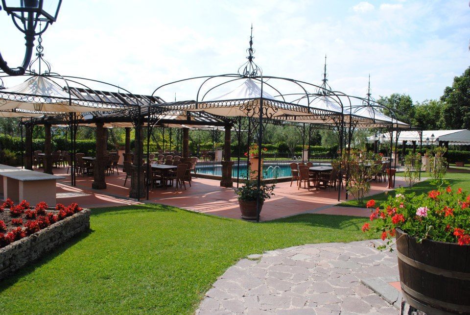 Zona pranzo all'aperto con tavoli sotto graticci decorati, piscina a vista, prato verde, patio in mattoni rossi.