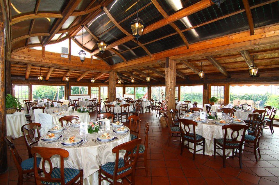 Sala da pranzo con tavoli rotondi apparecchiati per i pasti, travi e soffitto in legno, finestre con vegetazione visibile all'esterno.