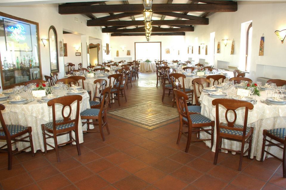 Sala da pranzo del ristorante con tavoli rotondi, sedie e centrotavola; pavimenti in cotto, pareti bianche e luce solare intensa.
