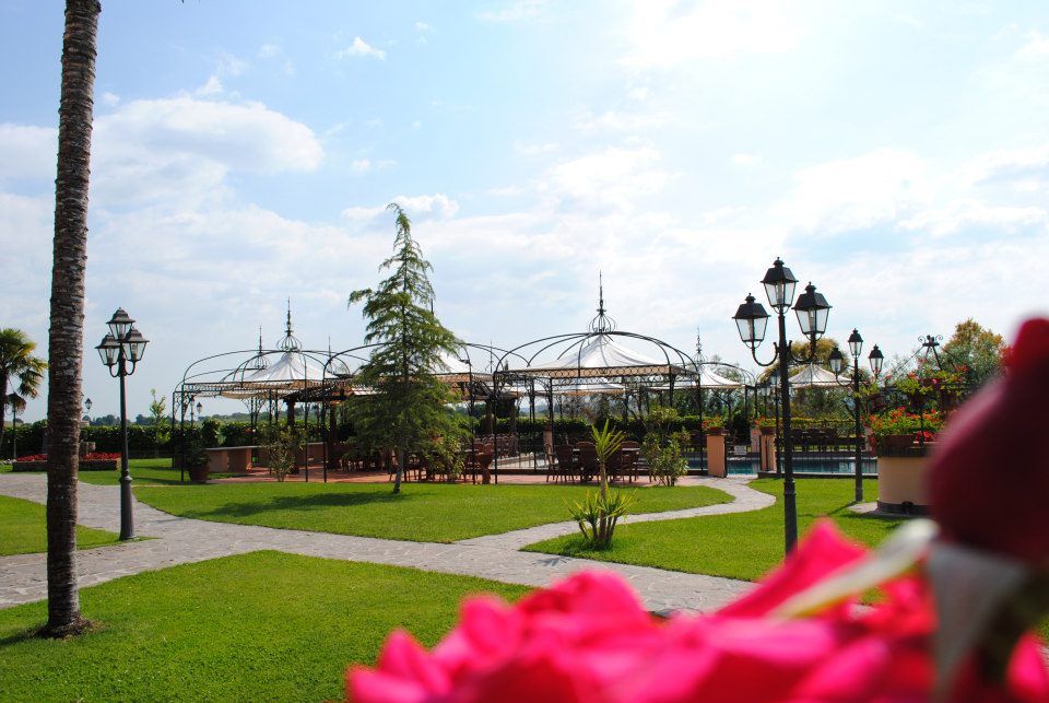 Prato verde, sentieri e zona pranzo all'aperto con tettoie bianche sotto un cielo azzurro brillante.