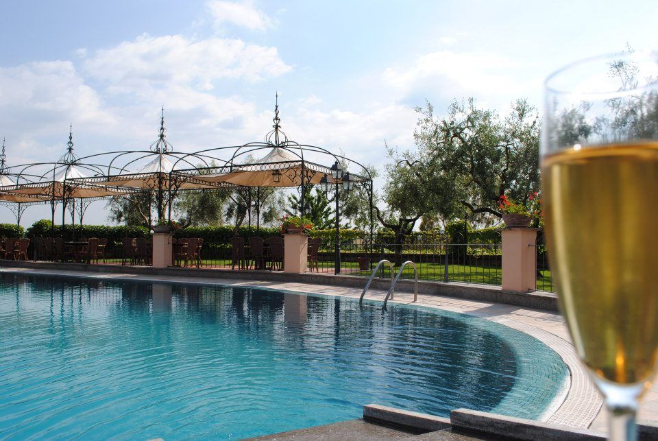 Piscina con bicchiere di champagne parzialmente visibile in primo piano, zona pranzo all'aperto e alberi sullo sfondo sotto un cielo azzurro.