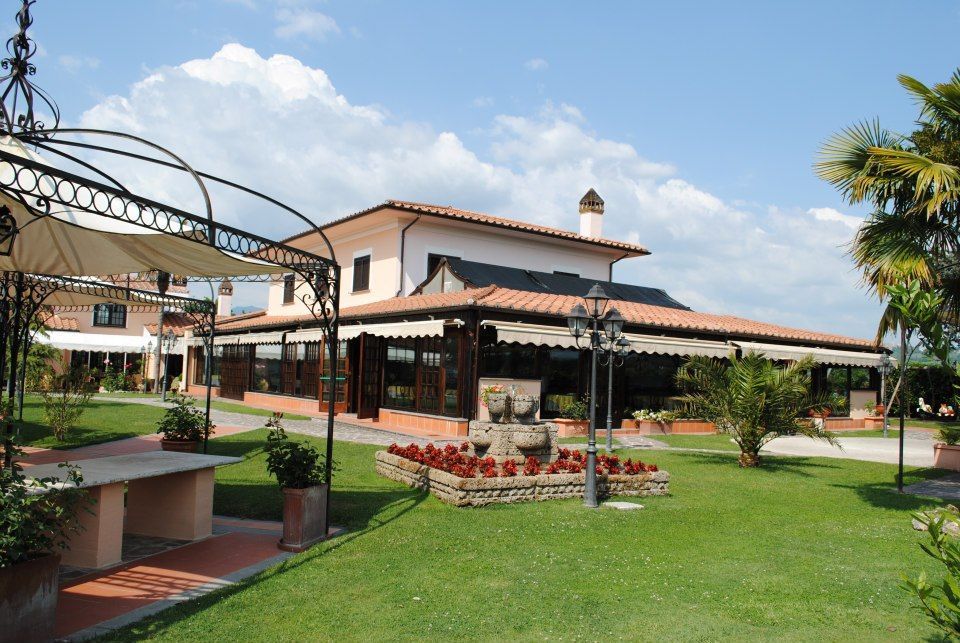 Ristorante con tetto di tegole rosse, ampie finestre e prato verde; gazebo e giardino fiorito in primo piano sotto un cielo azzurro brillante.