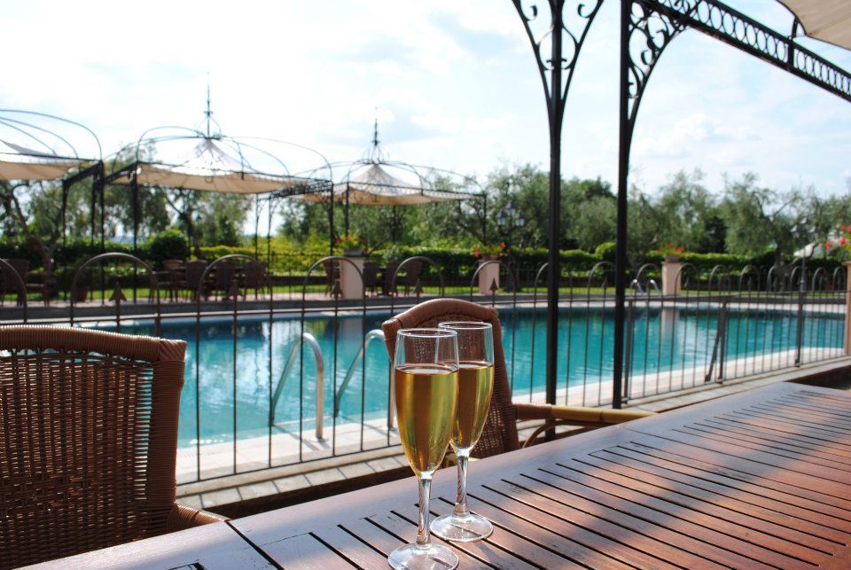 Due calici da champagne sul tavolo, con vista sulla piscina. Patio esterno con posti a sedere coperti e alberi.