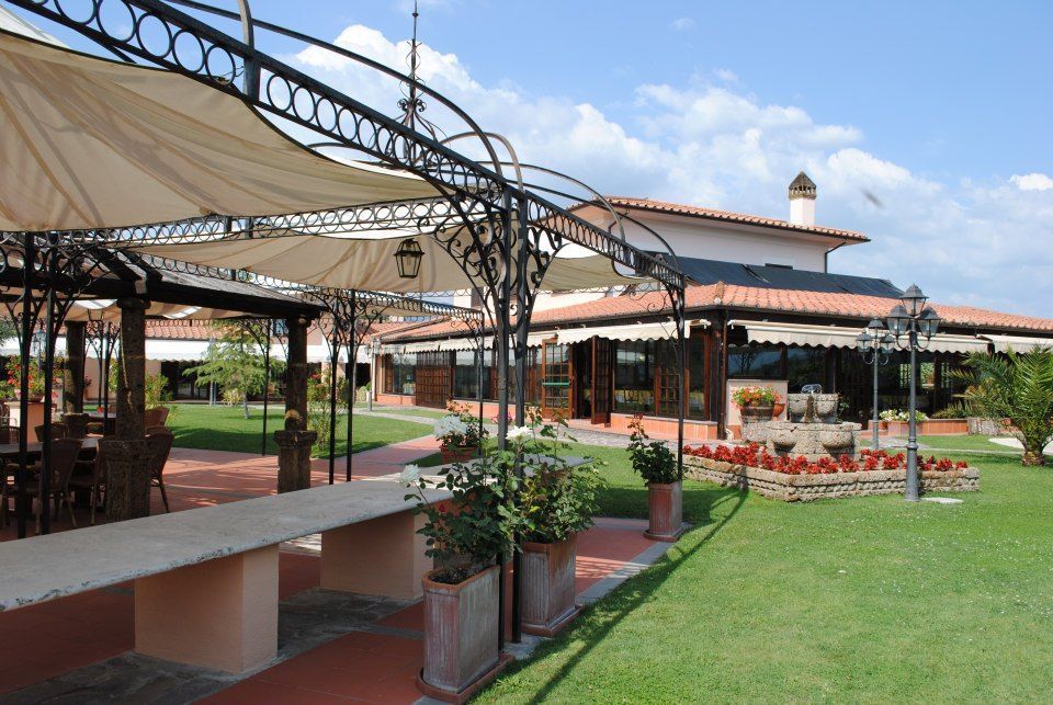 Ristorante all'aperto con tettoia in ferro battuto, tavoli e tetto di tegole rosse, su un prato erboso sotto un cielo soleggiato.