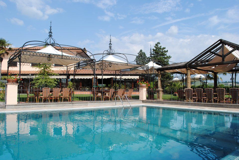 Zona pranzo a bordo piscina con tavoli, sedie e tende da sole sotto un cielo azzurro.