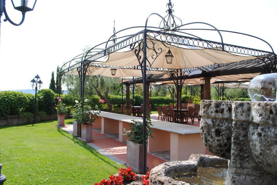 Zona pranzo all'aperto con pergolato rivestito in tessuto beige. Tavoli, sedie e fontana in pietra nelle giornate di sole.