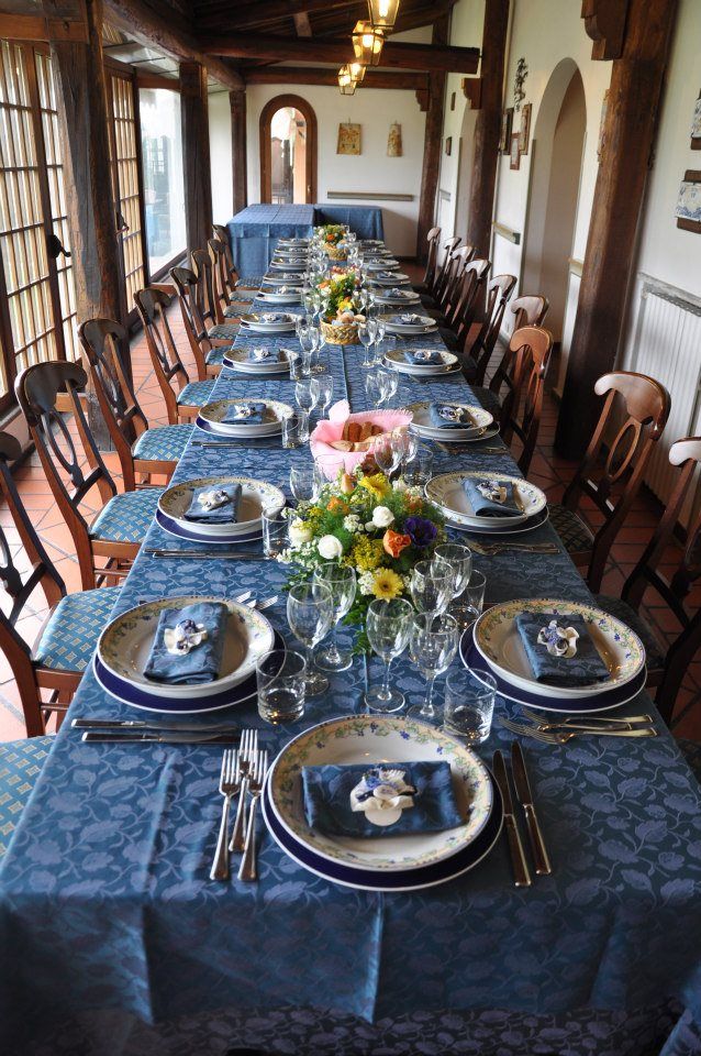 Lungo tavolo da pranzo apparecchiato per un pasto, tovaglia blu, centrotavola floreale, sedie e finestre.