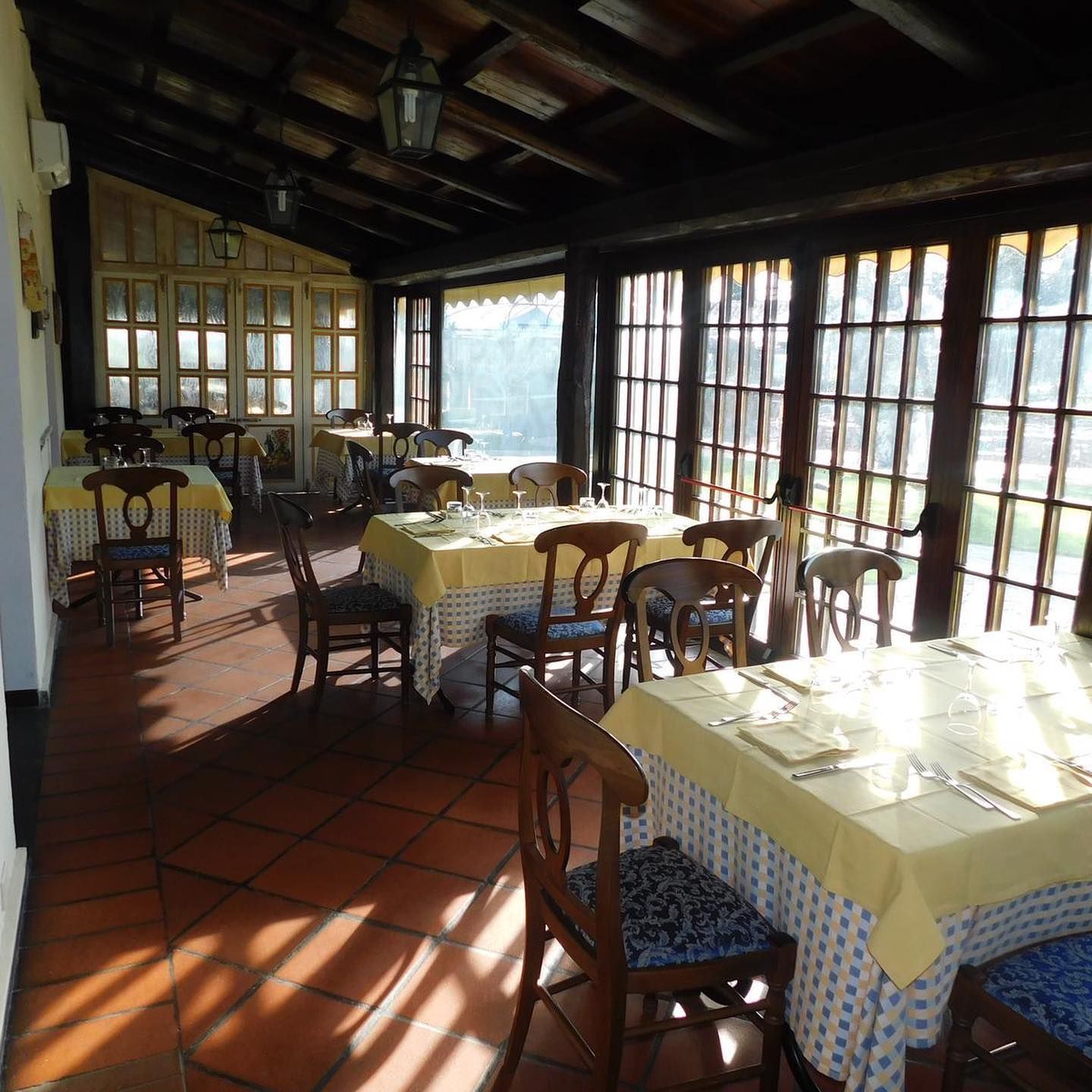 Interno del ristorante illuminato dal sole, con tavoli apparecchiati per cenare. Tovaglie gialle, sedie in legno e ampie finestre.