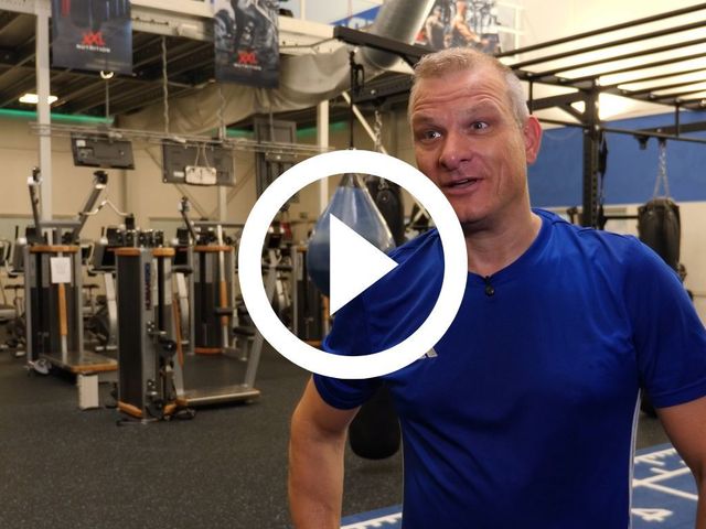 Man in blauw shirt in de sportschool, lachend. Fitnessapparatuur op de achtergrond.