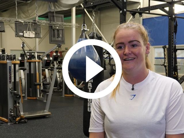 Een vrouw in een wit shirt lacht in een sportschool, met boksapparatuur en een bokszak op de achtergrond.