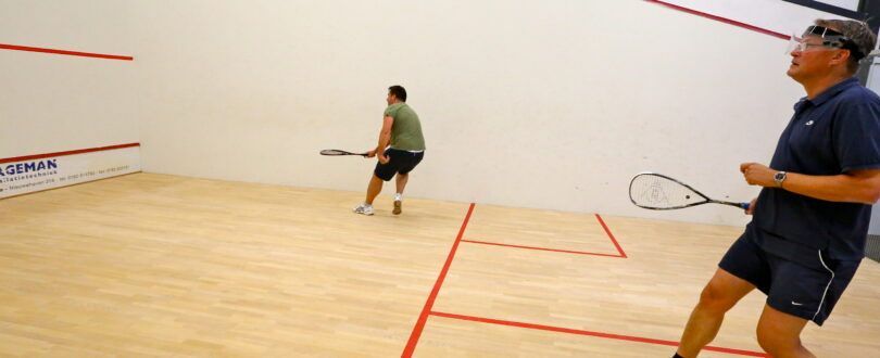 Twee mannen spelen squash op een veld. Eén man staat op de voorgrond met zijn racket en een andere op de achtergrond.
