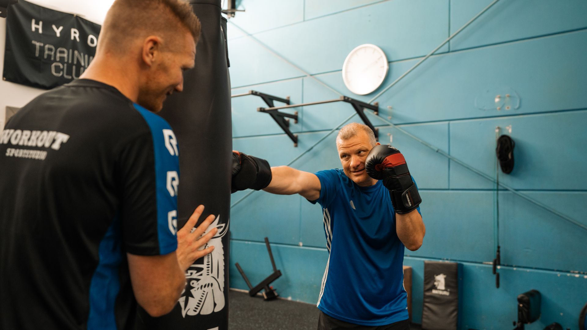 Man bokst met trainer in de sportschool. Blauwe en zwarte bokshandschoenen, zwarte bokszak, lichtblauwe muur.