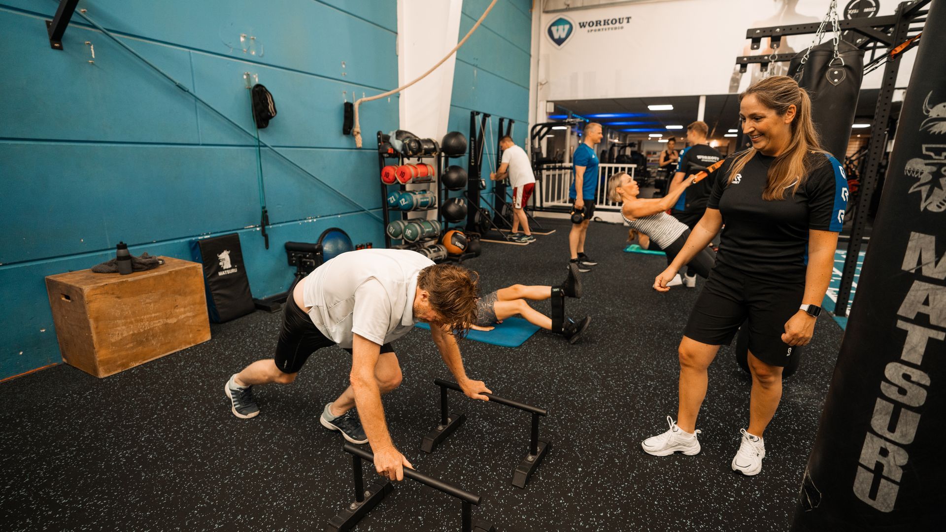 Een fitnessles in een sportschool. Een vrouw coacht een man bij het doen van een hordenoefening, terwijl de anderen trainen.