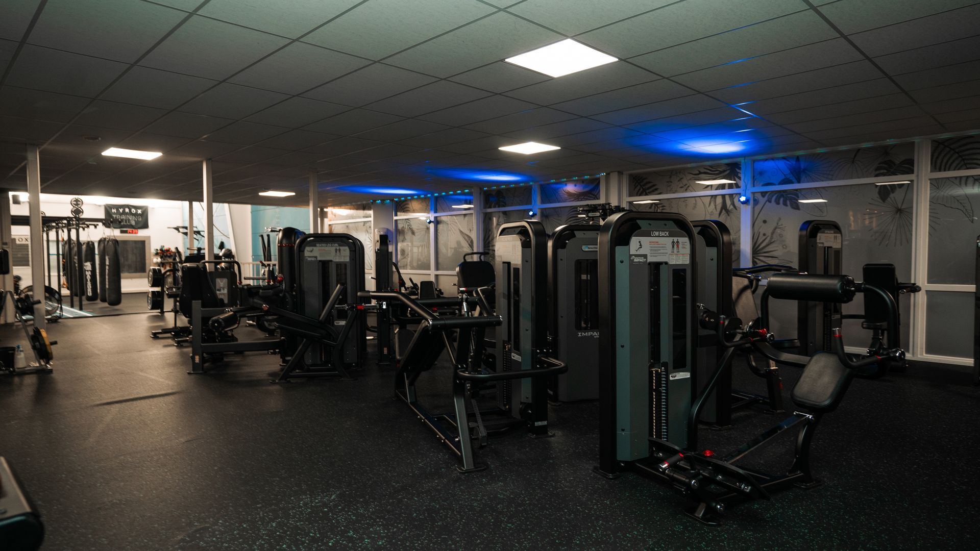 Interieur van een sportschool met fitnessapparaten, donkere vloer en blauwe verlichting.