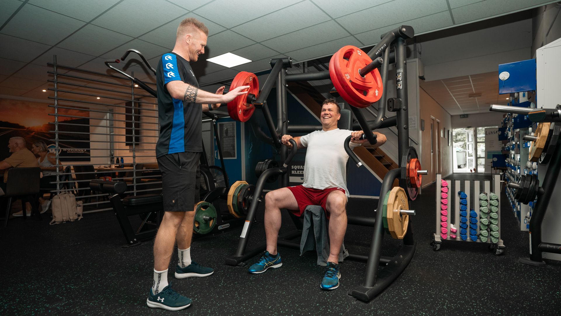Man using a weight machine, assisted by a trainer, in a gym.