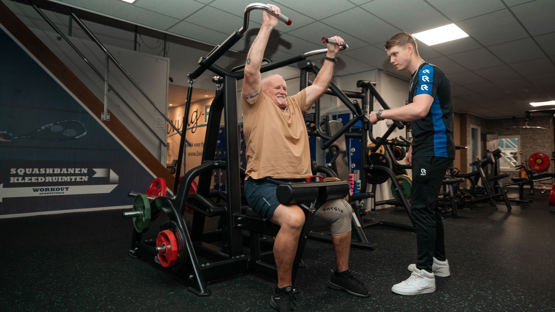 Oudere man gebruikt fitnessapparaat met hulp van trainer. In de sportschool draagt de man een kniebrace.