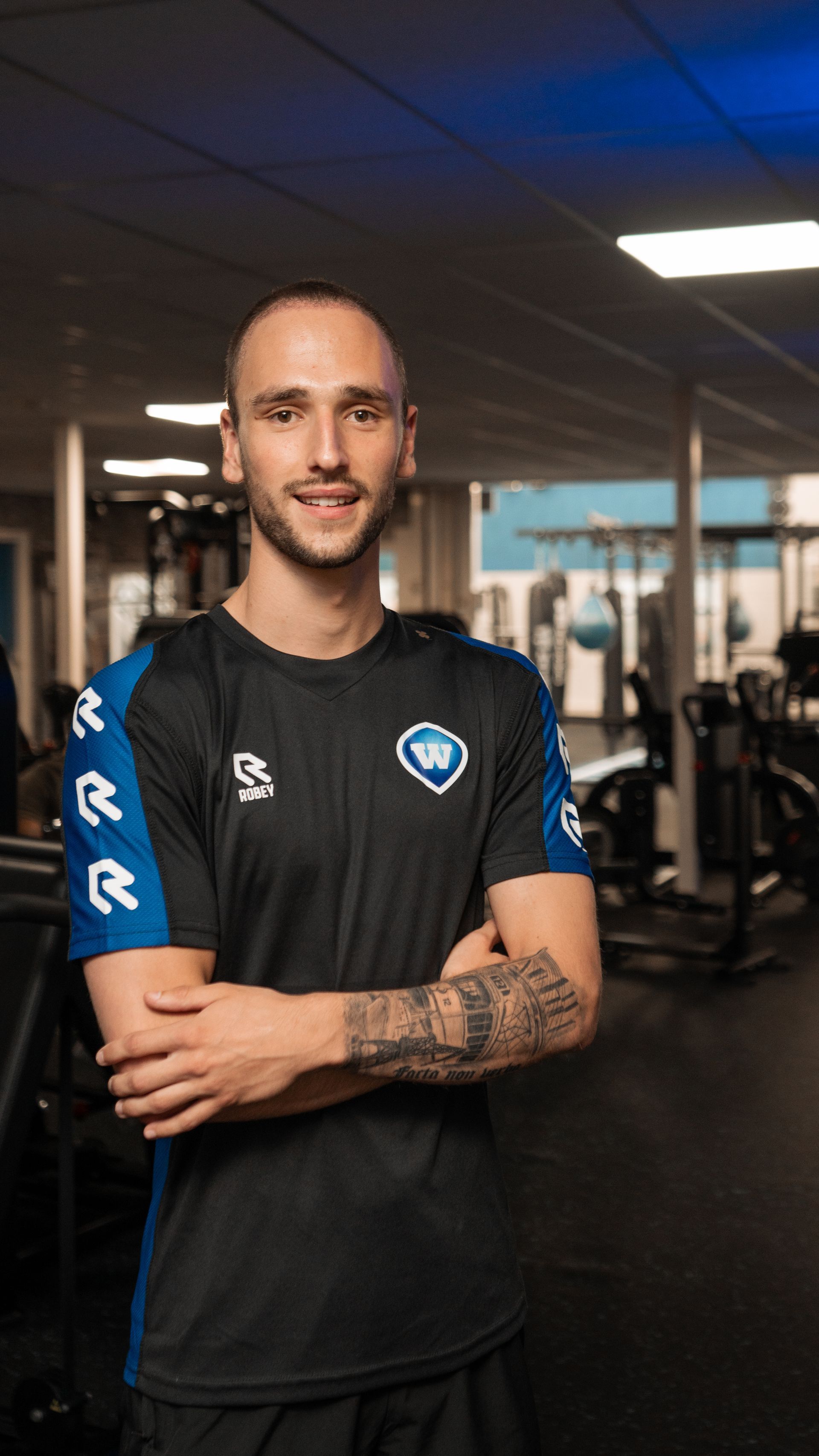 Man met tatoeages op zijn arm in de sportschool, gekleed in een zwart-blauw shirt, lachend en met gekruiste armen.