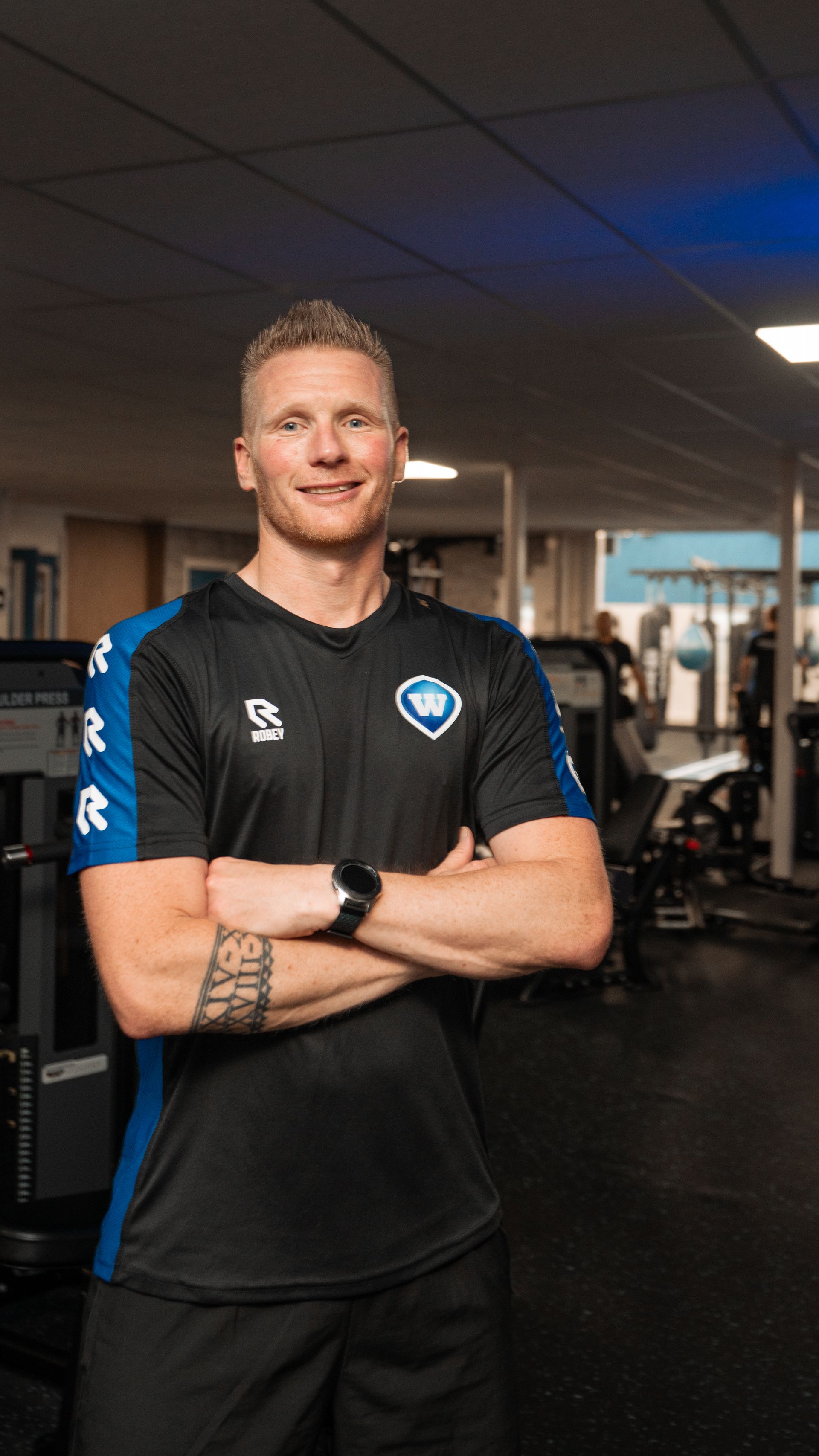 Man met gekruiste armen in een sportschool, gekleed in een zwart shirt met blauwe accenten, glimlachend.