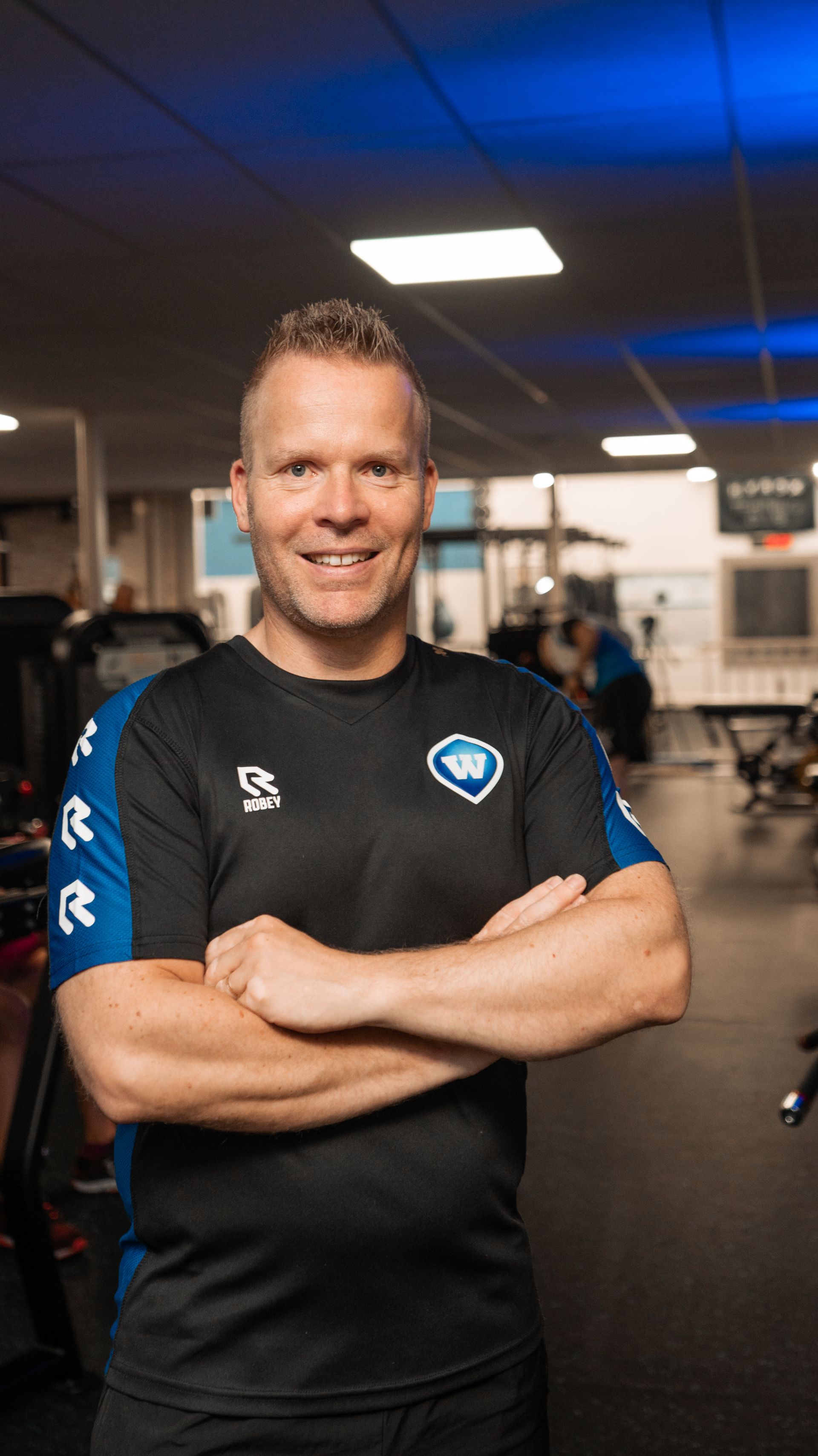 Man met gekruiste armen, lachend in een sportschool, gekleed in een zwart-blauw sportshirt.