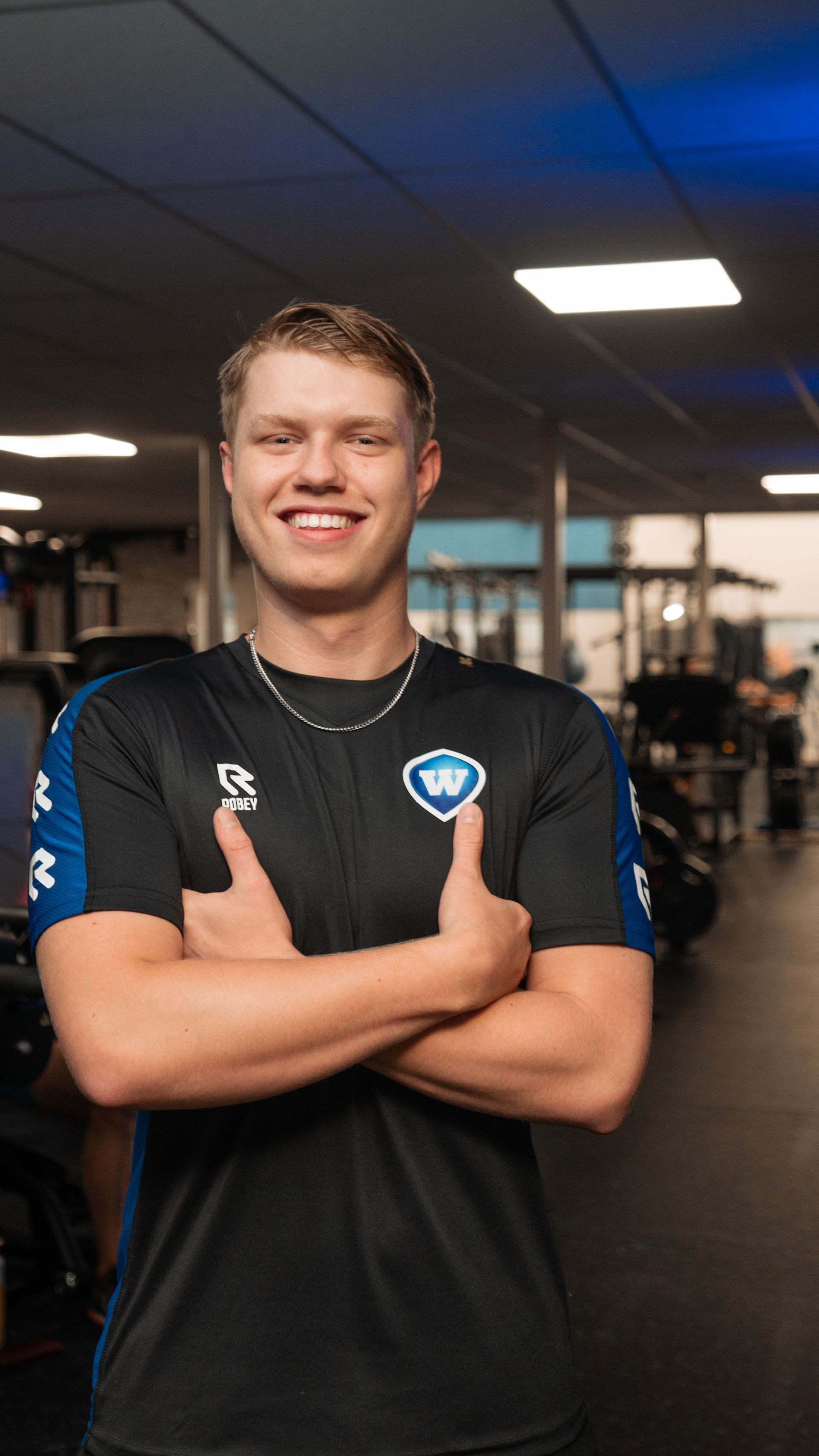 Man in sportschool met gekruiste armen, gekleed in een blauw en zwart shirt, lachend.