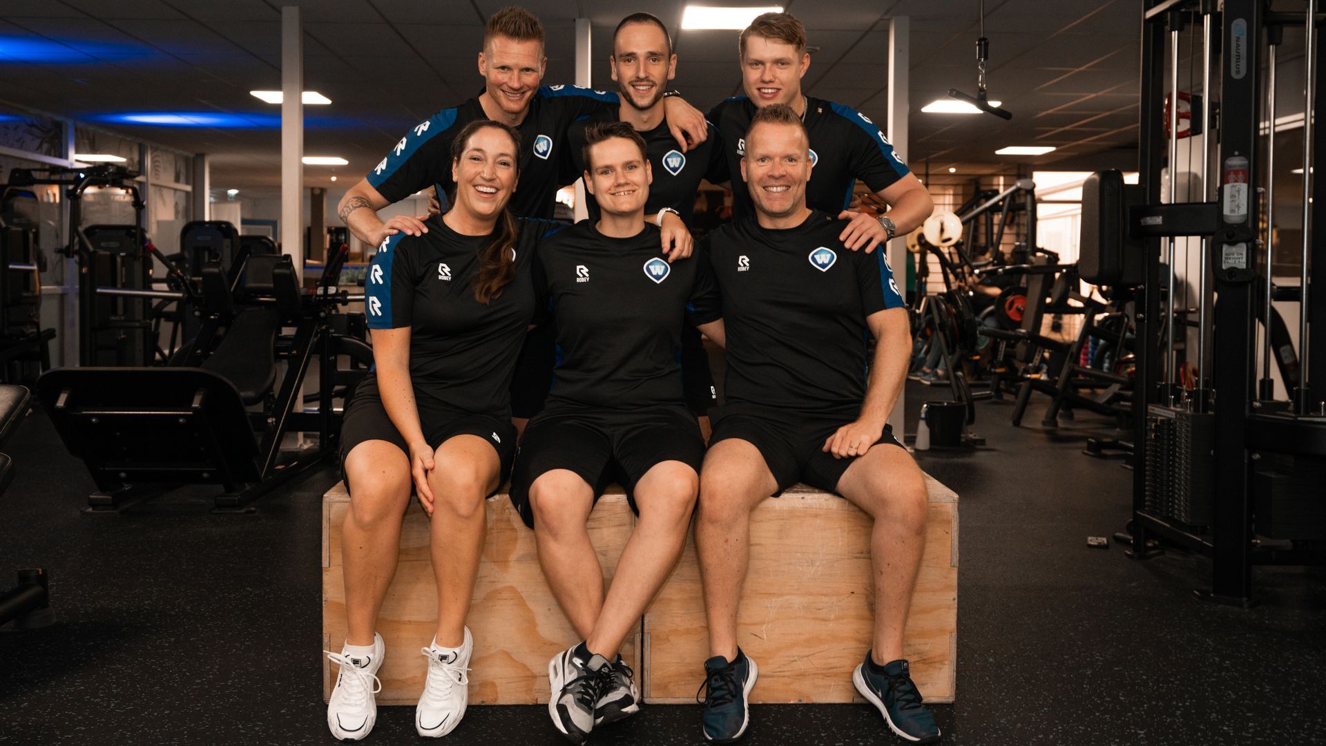 Vrouw helpt man met push-ups in sportschool, draagt sportkleding; rode opdrukstangen.